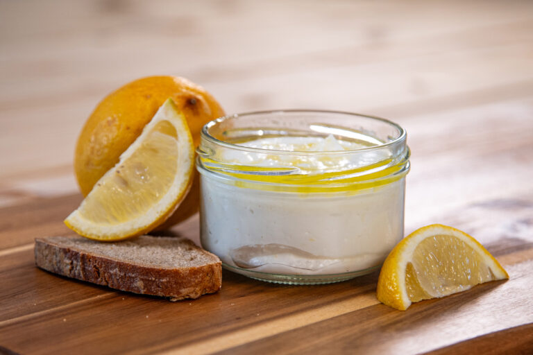 Produktfoto von Frischkäse Chevre au Citron in einem Glas auf einer Holzplatte mit Zitronenspalten und Brot dekoriert.