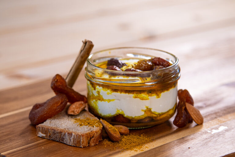 Produktfoto von Frischkäse Curry-Aprikose in einem Glas auf einer Holzplatte mit Curry, Mandeln, Zimt, getrockneten Aprikosen und Brot dekoriert.