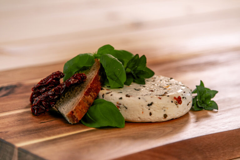 Produktfoto von Grillkäse mediterran auf einer Holzplatte mit getrockneten Tomaten, Basilikum und Brot dekoriert.
