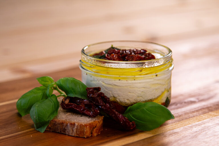 Produktfoto von Frischkäse Tomate-Basilikum in einem Glas auf einer Holzplatte mit getrockneten Tomaten, Basilikum und Brot dekoriert.