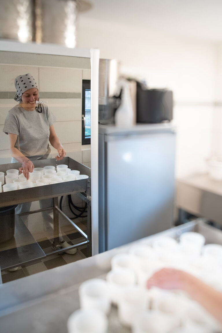 Spiegelung in einer Kühlschranktür von einer Frau mit Tuch auf dem Kopf die vor einem Tisch mit Plastikbehältern steht.