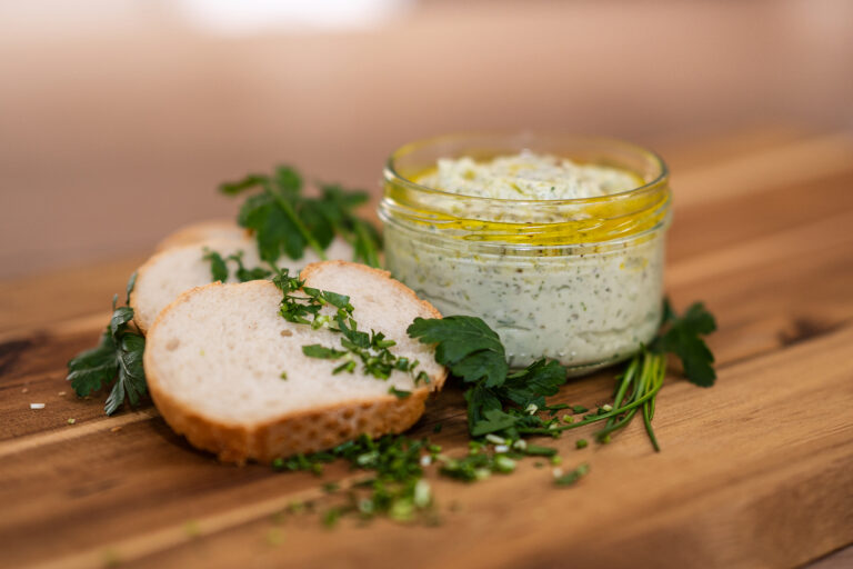 Dekoratives Foto von Ziegenfrischkäse in einem Glas auf einem Holzbrett zwischen Brotscheiben und Kräutern